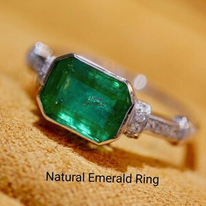 May include: A silver ring with a large emerald green gemstone and smaller diamonds set in the band. The ring is on a brown suede surface. Natural Emerald Ring.
