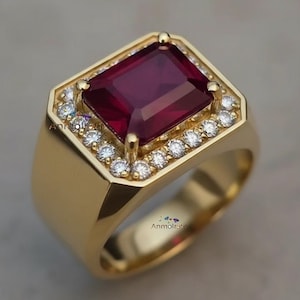 May include: A gold ring featuring a large, square-cut, deep red gemstone, surrounded by a halo of small, sparkling white stones. The ring has a classic, rectangular design with a polished finish, showcasing a luxurious aesthetic.