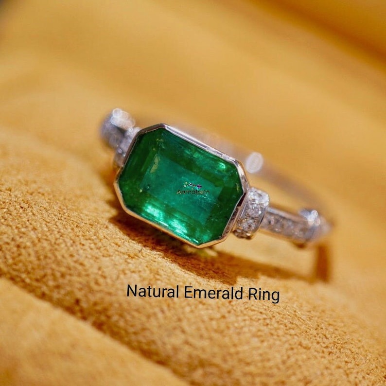 May include: A silver ring with a large emerald green gemstone and smaller diamonds set in the band. The ring is on a brown suede surface. Natural Emerald Ring.