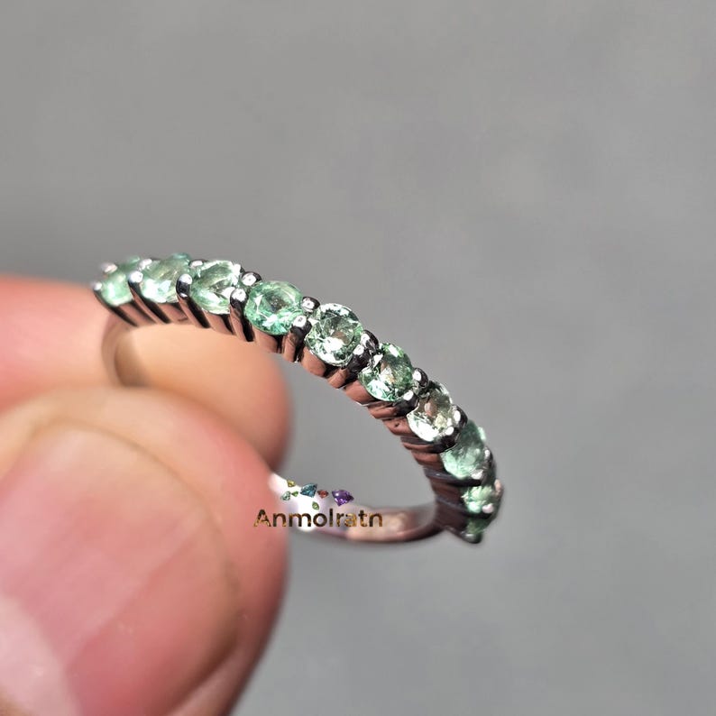 May include: A silver ring featuring a row of round, light green gemstones. The ring is held by a hand against a gray background. The text "Anmolratn" is visible on the ring.