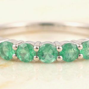 May include: Close-up of a silver ring set with multiple round, vibrant green emeralds. The gemstones are evenly spaced and securely set in the band, showcasing their color and brilliance. The ring's design is simple, elegant, and suitable for everyday wear.