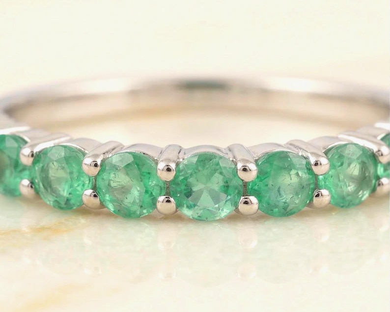 May include: Close-up of a silver ring set with multiple round, vibrant green emeralds. The gemstones are evenly spaced and securely set in the band, showcasing their color and brilliance. The ring's design is simple, elegant, and suitable for everyday wear.