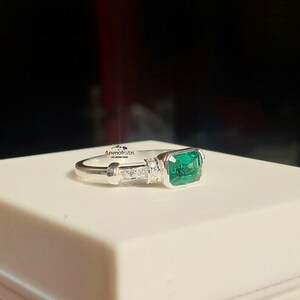 May include: A silver ring with a large emerald green gemstone and smaller clear gemstones set in the band. The ring is on a white surface.