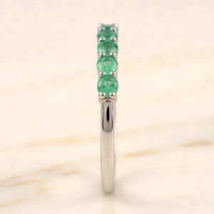 May include: A silver ring featuring a row of round, vibrant green emeralds. The gemstones are set closely together, creating a continuous line of color. The band is smooth and polished, reflecting light.
