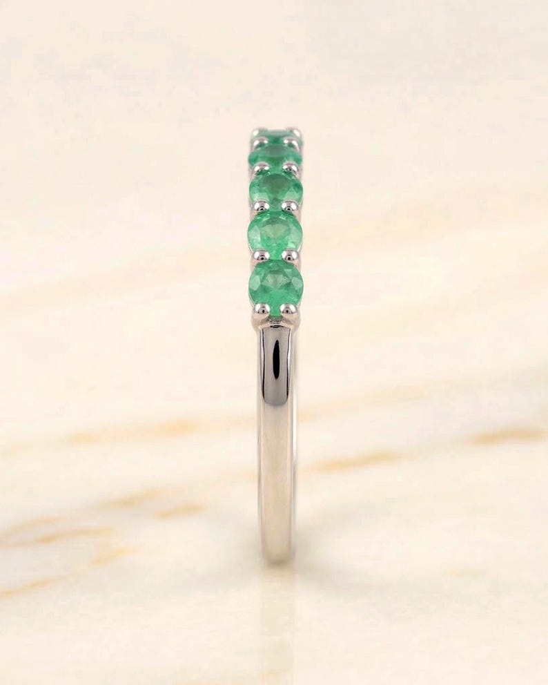May include: A silver ring featuring a row of round, vibrant green emeralds. The gemstones are set closely together, creating a continuous line of color. The band is smooth and polished, reflecting light.