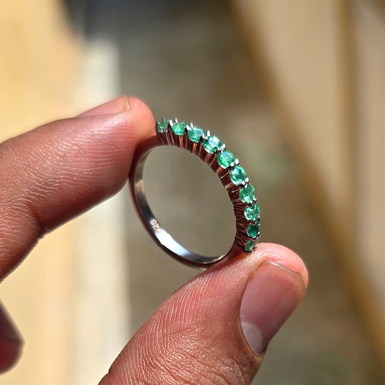 May include: A silver ring with a row of small, round, bright green emeralds. The ring is held up, showing the gemstones and the polished metal band. The ring is a piece of jewelry.
