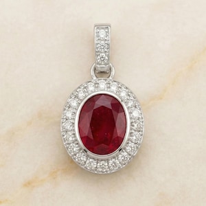 May include: Oval pendant featuring a vibrant red ruby encircled by a halo of small diamonds. The pendant is set in white metal, possibly platinum or white gold, with a diamond-set bale for a chain. The ruby is the focal point.