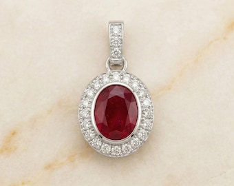 Puede incluir: Colgante ovalado con una gran piedra central de rubí rojo intenso, rodeada por un halo de pequeños diamantes brillantes. El colgante está engastado en un metal blanco, probablemente platino u oro blanco, y tiene una fianza con diamantes.