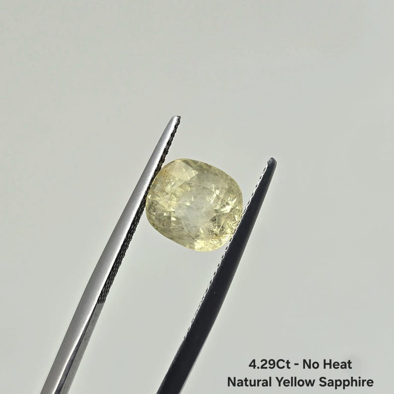 Op de afbeelding: Een natuurlijke gele saffier in kussenslijpvorm, vastgehouden door een metalen pincet. De steen is lichtgeel met zichtbare interne facetten. De tekst op de afbeelding luidt: 4.29Ct - No Heat Natural Yellow Sapphire. De edelsteen is doorschijnend.