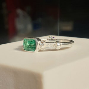 May include: A silver ring with a large emerald green gemstone set in a square bezel. The ring has a band of small diamonds set in a row.