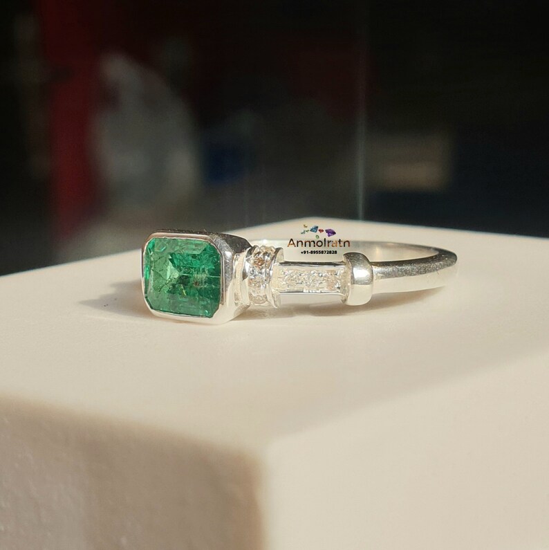 May include: A silver ring with a large emerald green gemstone set in a square bezel. The ring has a band of small diamonds set in a row.