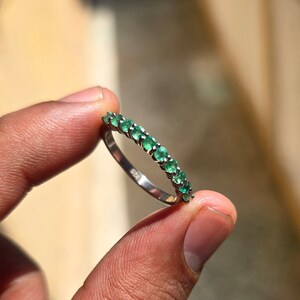 May include: A silver ring with a row of vibrant green emeralds. The ring is held between two fingers, showcasing the polished metal and the sparkling gemstones. The ring is a delicate piece of jewelry.