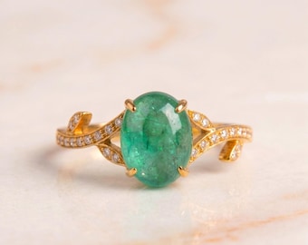 May include: A gold ring featuring a large, oval-shaped, cabochon-cut green emerald center stone held by four prongs. The band is designed with a split-shank leaf motif, accented with small, round, clear pavé-set diamonds along the sides.