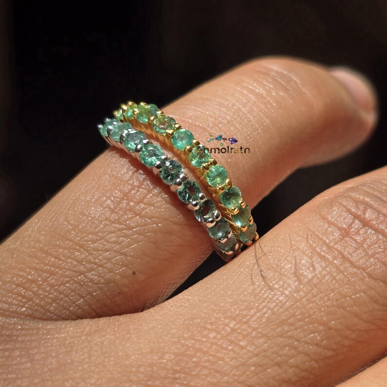 May include: Two gemstone rings, one gold and one silver, each adorned with a row of small, round emeralds. The rings are stacked on a finger, showcasing the vibrant green and teal hues of the gemstones.