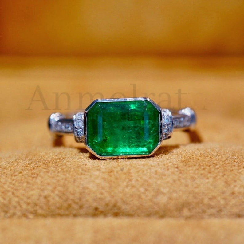 May include: A silver ring with a large emerald green gemstone surrounded by smaller diamonds. The ring is set on a brown background.