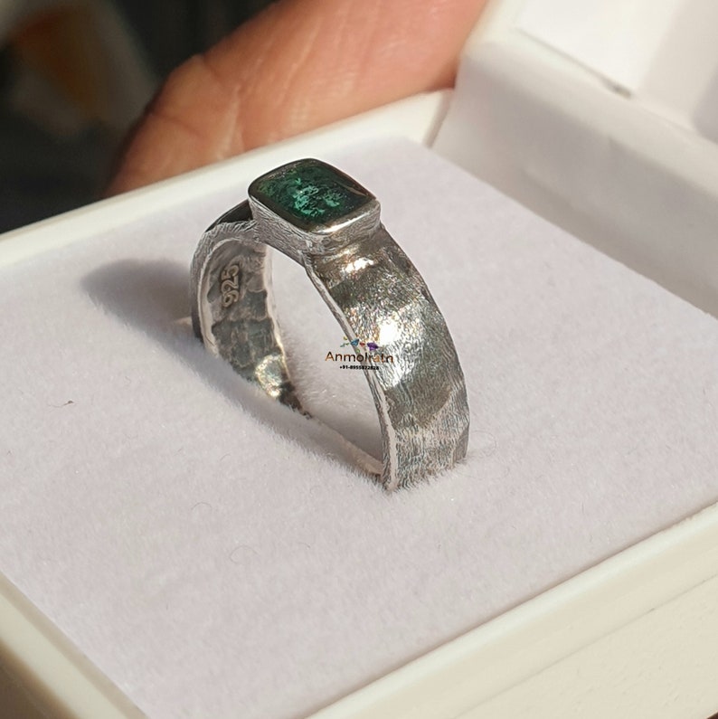K&ouml;nnte beinhalten: Ein silberner Ring mit geh&auml;mmerter Oberfl&auml;che und einem gr&uuml;nen Edelstein, der in einer quadratischen Fassung sitzt. Der Ring befindet sich in einer wei&szlig;en Ringbox.