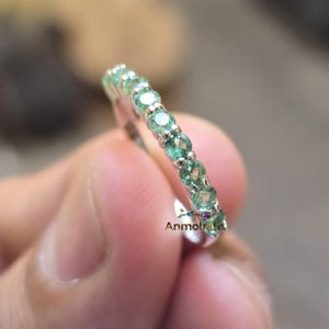 May include: A silver ring with a row of round, light green gemstones. The ring is held in a person's hand, with the gemstones catching the light. The ring is a piece of jewelry.