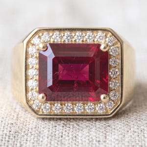 Könnte beinhalten: Ein Goldring mit einem großen, rechteckigen, tiefroten Edelstein in der Mitte. Der zentrale Stein ist von einem Kranz aus kleinen, runden, klaren weißen Diamanten umgeben, die in das Goldband mit glatter, polierter Oberfläche gefasst sind.