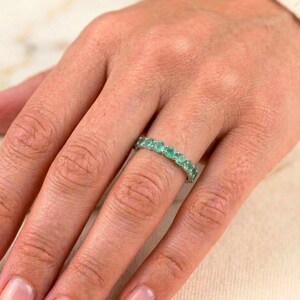 May include: A silver ring with a row of oval-cut emeralds. The ring is worn on a finger, showcasing the vibrant green gemstones. The ring's design is simple, highlighting the beauty of the emeralds.