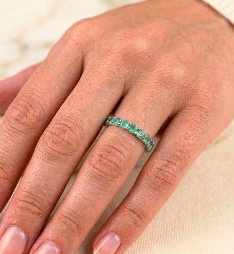 May include: A silver ring with a row of oval-cut emeralds. The ring is worn on a finger, showcasing the vibrant green gemstones. The ring's design is simple, highlighting the beauty of the emeralds.