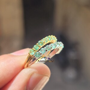 May include: Two gemstone rings, one gold and one silver, each set with a row of small, round, green-blue gemstones. The gold ring has a warmer, yellowish hue, while the silver ring has a cooler, brighter appearance. The rings are held up against a blurred background.