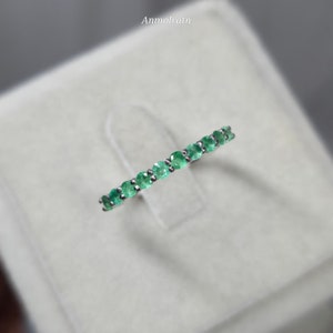 May include: A silver ring featuring a row of vibrant green emeralds. The gemstones are set closely together, creating a continuous band of color. The ring is displayed on a soft, neutral background, highlighting the sparkle of the emeralds.