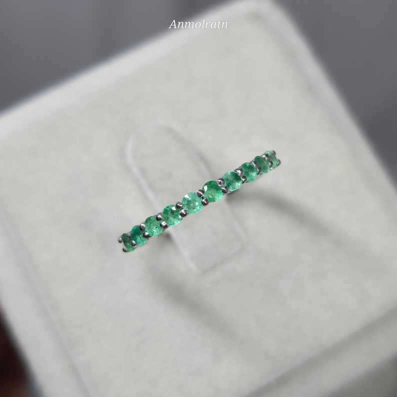 May include: A silver ring featuring a row of vibrant green emeralds. The gemstones are set closely together, creating a continuous band of color. The ring is displayed on a soft, neutral background, highlighting the sparkle of the emeralds.