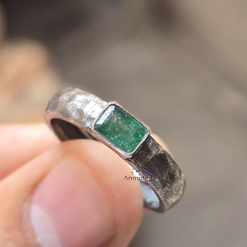 K&ouml;nnte beinhalten: Ein silberner Ring mit einer strukturierten Band und einem kleinen, rechteckigen smaragdgr&uuml;nen Edelstein, der in einer silbernen Fassung sitzt.