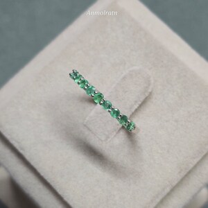 May include: A delicate ring featuring a row of vibrant green emeralds set in a silver band. The gemstones are round-cut and evenly spaced, creating a continuous line of color. The ring is displayed on a neutral-toned surface.