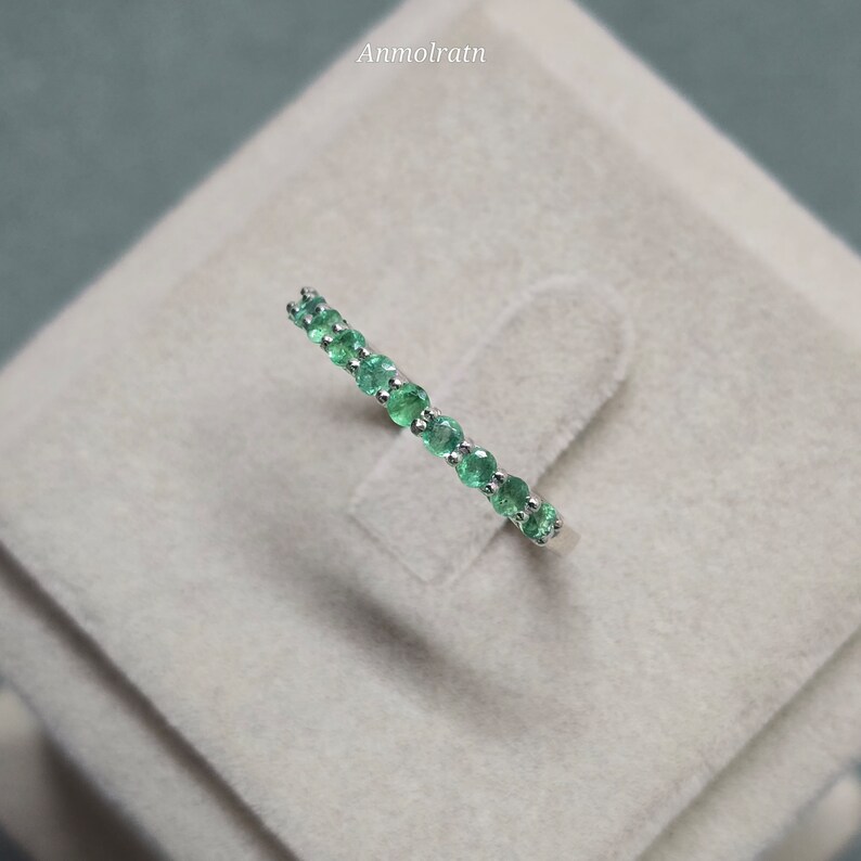 May include: A delicate ring featuring a row of vibrant green emeralds set in a silver band. The gemstones are round-cut and evenly spaced, creating a continuous line of color. The ring is displayed on a neutral-toned surface.