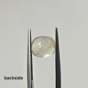 Op de afbeelding: Een gefacetteerde, lichtgele, ovaalvormige edelsteen vastgehouden door een metalen pincet. De steen heeft een doorschijnend uiterlijk met zichtbare interne insluitingen en een gepolijst oppervlak. Het woord "backside" staat in zwarte letters linksonder.