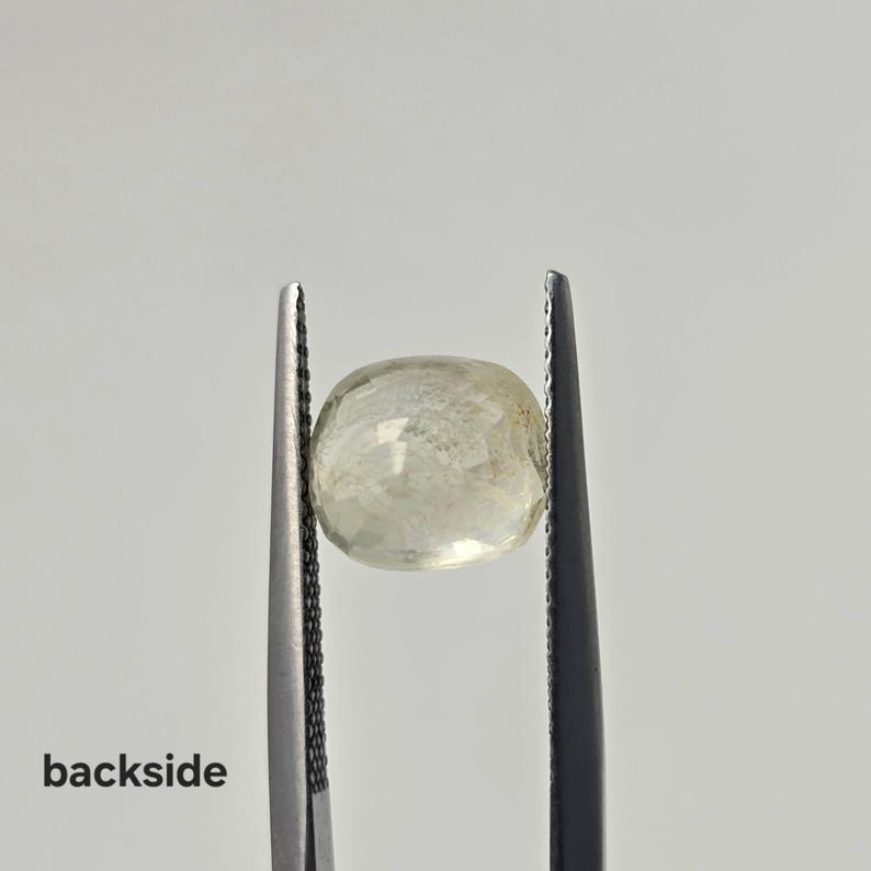 Op de afbeelding: Een gefacetteerde, lichtgele, ovaalvormige edelsteen vastgehouden door een metalen pincet. De steen heeft een doorschijnend uiterlijk met zichtbare interne insluitingen en een gepolijst oppervlak. Het woord "backside" staat in zwarte letters linksonder.