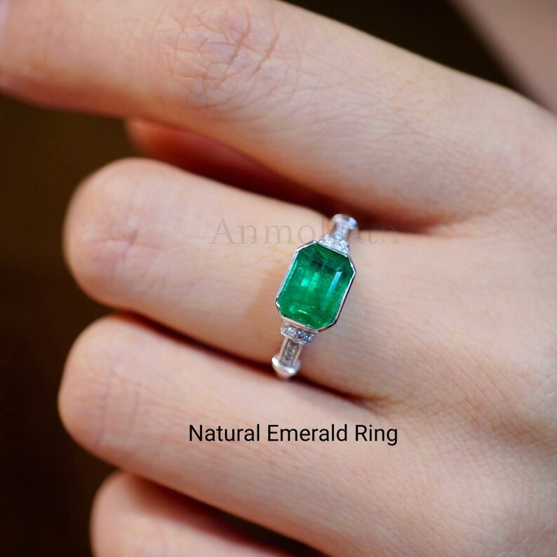 May include: A silver ring with a large emerald green gemstone set in a square shape. The ring is accented with smaller diamonds on the band. "Natural Emerald Ring" is written below the ring.