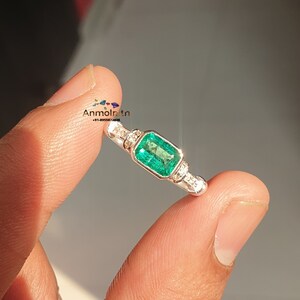 May include: A silver ring with a large emerald green gemstone surrounded by smaller diamonds. The ring is on a finger.