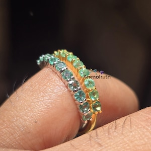 May include: Two rings with a row of small, round gemstones. One ring has silver-colored metal and teal-colored stones. The other ring has gold-colored metal and green stones. The rings are on a finger.