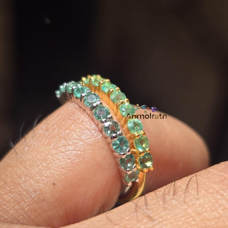 May include: Two rings with a row of small, round gemstones. One ring has silver-colored metal and teal-colored stones. The other ring has gold-colored metal and green stones. The rings are on a finger.