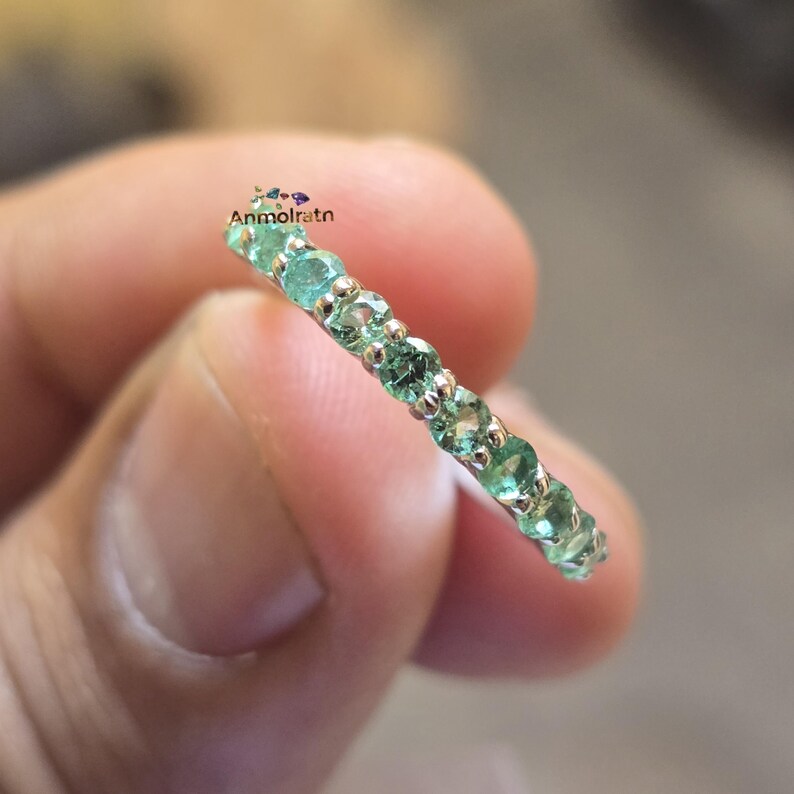 May include: A silver ring with a row of small, round, light green gemstones. The ring is held in a person's hand, and the gemstones are evenly spaced around the band. The ring is a delicate piece of jewelry.