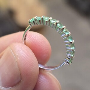 May include: A silver ring featuring a row of oval-cut emeralds. The ring's band is thin and polished, with the gemstones set closely together. The emeralds have a vibrant green color, and the ring is held by a person's fingers.