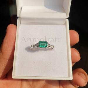 May include: A white gold ring with a large emerald green gemstone and smaller diamonds on the band. The ring is in a white box with the word "Anmolratn" printed on the inside.