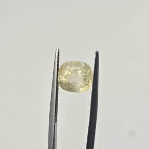 Op de afbeelding: Een lichtgele edelsteen in kussenslijpvorm, vastgehouden door een donkere metalen pincet. De steen heeft een gefacetteerd oppervlak dat licht reflecteert en vertoont een doorschijnende kwaliteit met interne insluitingen in het heldere, lichtgele lichaam.