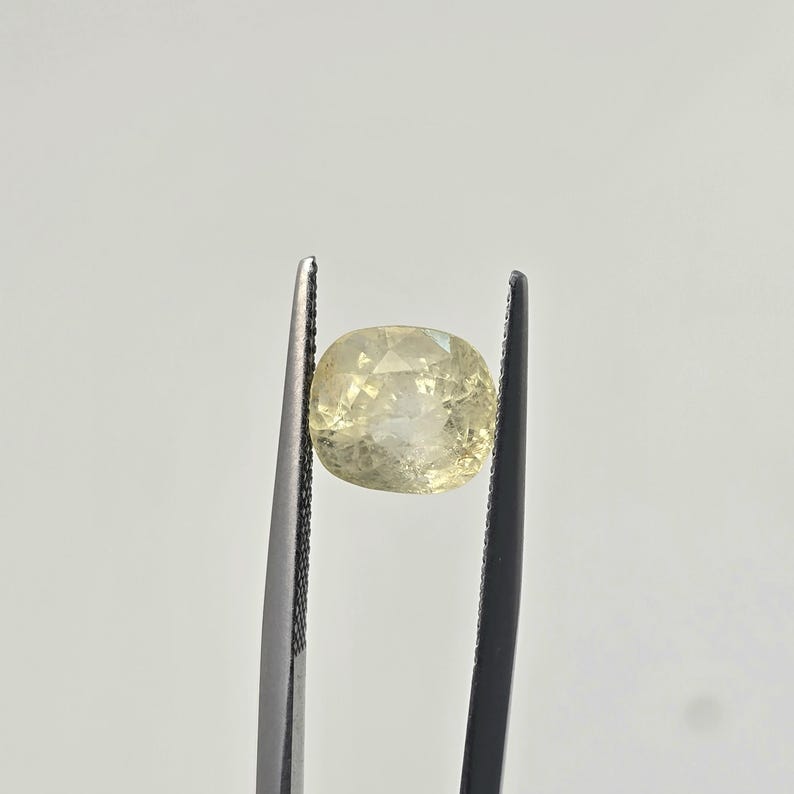 Op de afbeelding: Een lichtgele edelsteen in kussenslijpvorm, vastgehouden door een donkere metalen pincet. De steen heeft een gefacetteerd oppervlak dat licht reflecteert en vertoont een doorschijnende kwaliteit met interne insluitingen in het heldere, lichtgele lichaam.