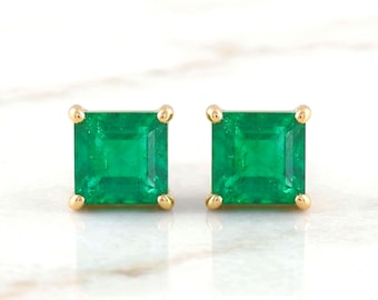May include: A pair of square emerald stud earrings with gold settings. The vibrant green gemstones are set in a four-prong design, showcasing their clarity and brilliance. The earrings are displayed on a white surface.