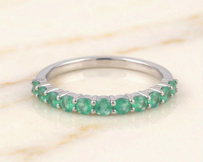 May include: A silver ring with a row of vibrant green emeralds. The gemstones are round-cut and set closely together, creating a continuous band of color. The ring is displayed on a light-colored surface.