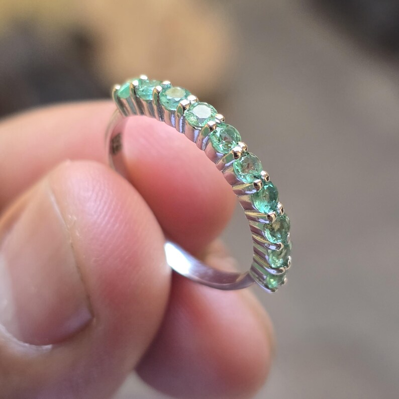 May include: A silver ring featuring a row of small, round, light green gemstones. The gemstones are evenly spaced and set in a silver band. The ring is held in a person's hand, with a blurred background.