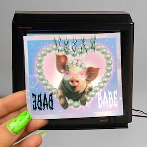 May include: A sticker featuring a cartoon pig wearing sunglasses and a crown. The pig is surrounded by a pearl necklace and the words "VEGAN BABE" are written on the sticker.