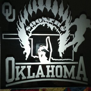 Op de afbeelding: Een zwart-witte afbeelding van een Indiaanse hoofdtooi met het woord "Sooners" erin. De afbeelding bevat ook een basketbalring en het woord "Oklahoma" eronder.