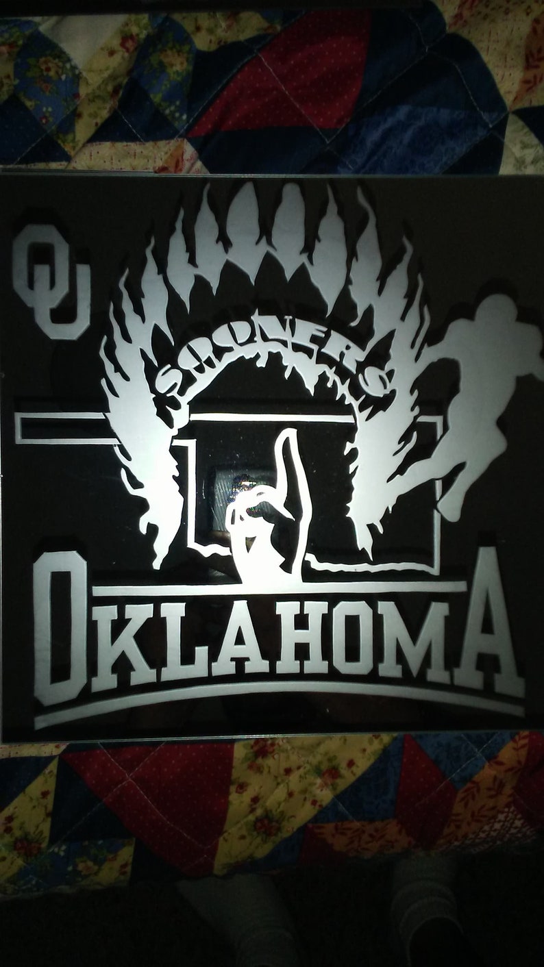 Op de afbeelding: Een zwart-witte afbeelding van een Indiaanse hoofdtooi met het woord "Sooners" erin. De afbeelding bevat ook een basketbalring en het woord "Oklahoma" eronder.