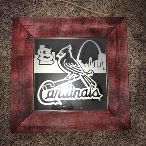 Op de afbeelding: Een ingelijst kunstwerk met een rode kardinaal met een honkbalknuppel, het logo van de St. Louis Cardinals en de Gateway Arch. De lijst is van verweerd rood hout.