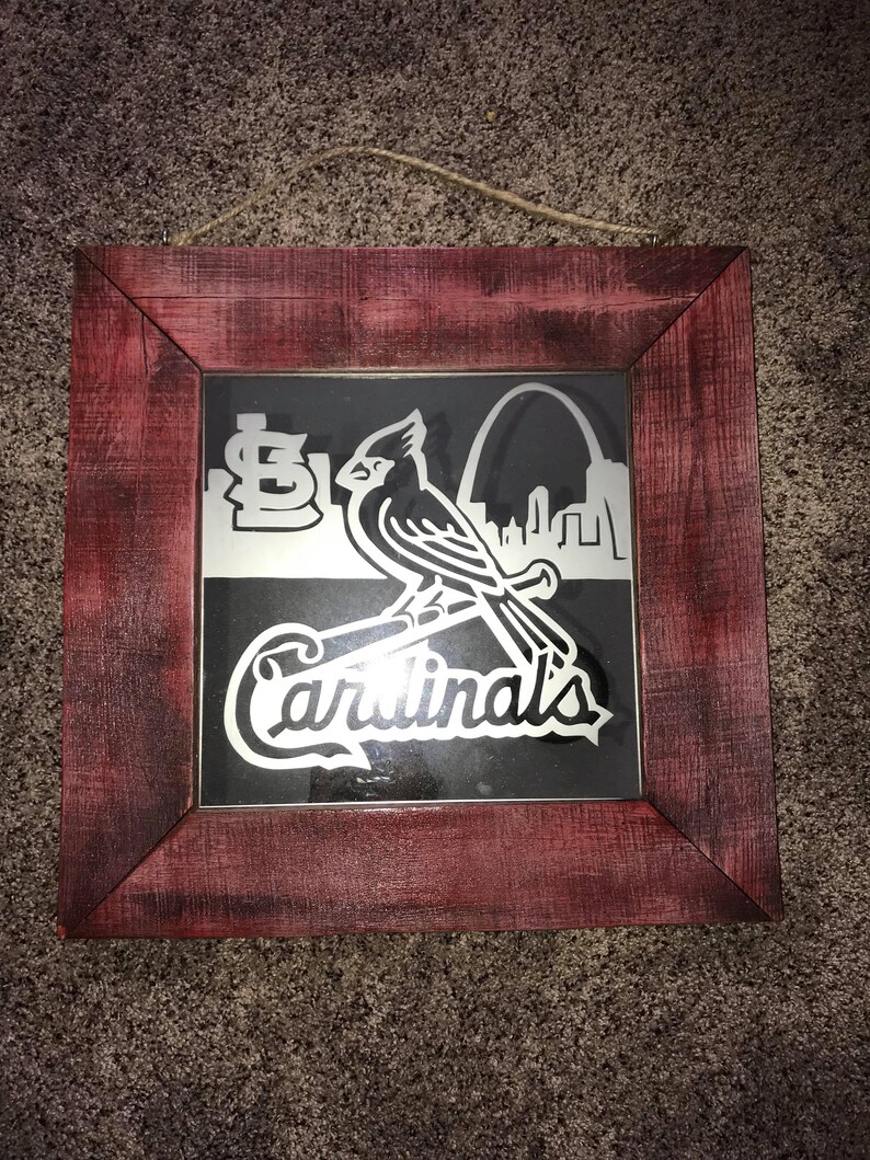 Op de afbeelding: Een ingelijst kunstwerk met een rode kardinaal met een honkbalknuppel, het logo van de St. Louis Cardinals en de Gateway Arch. De lijst is van verweerd rood hout.