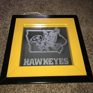 Op de afbeelding: Een ingelijste spiegel met een zwart en geel frame. De spiegel heeft een grijs silhouet van een havik die over de staat Iowa vliegt met het woord "HAWKEYES" eronder.